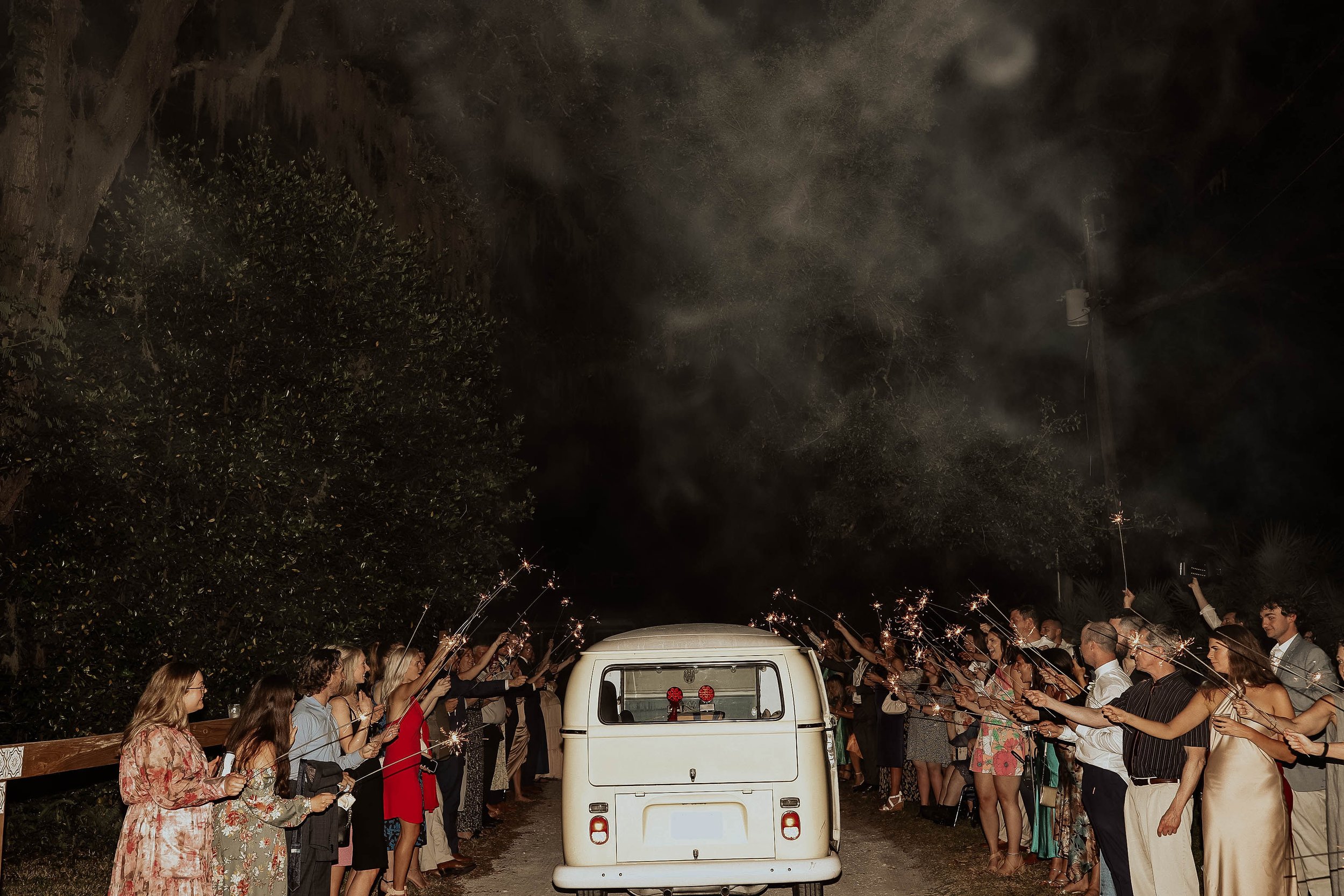 People celebrating with sparklers in a line, forming a tunnel, at night with trees in the background and a small white vehicle in the center.
