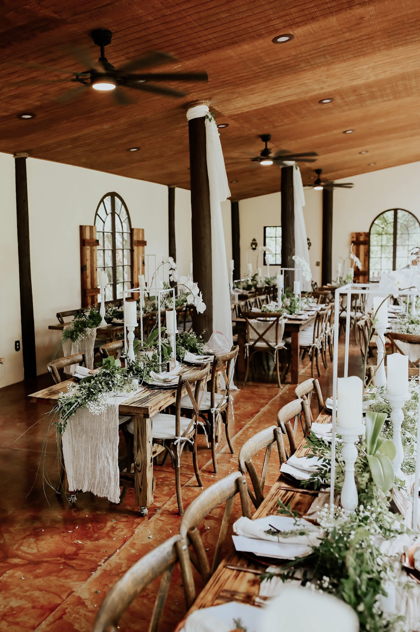Elegant rustic wedding reception setup with long wooden tables decorated with white candles, green foliage, and white flowers. Chairs surround the tables, and a warm wooden ceiling with ceiling fans is visible. Large windows with wooden accents provide natural light.