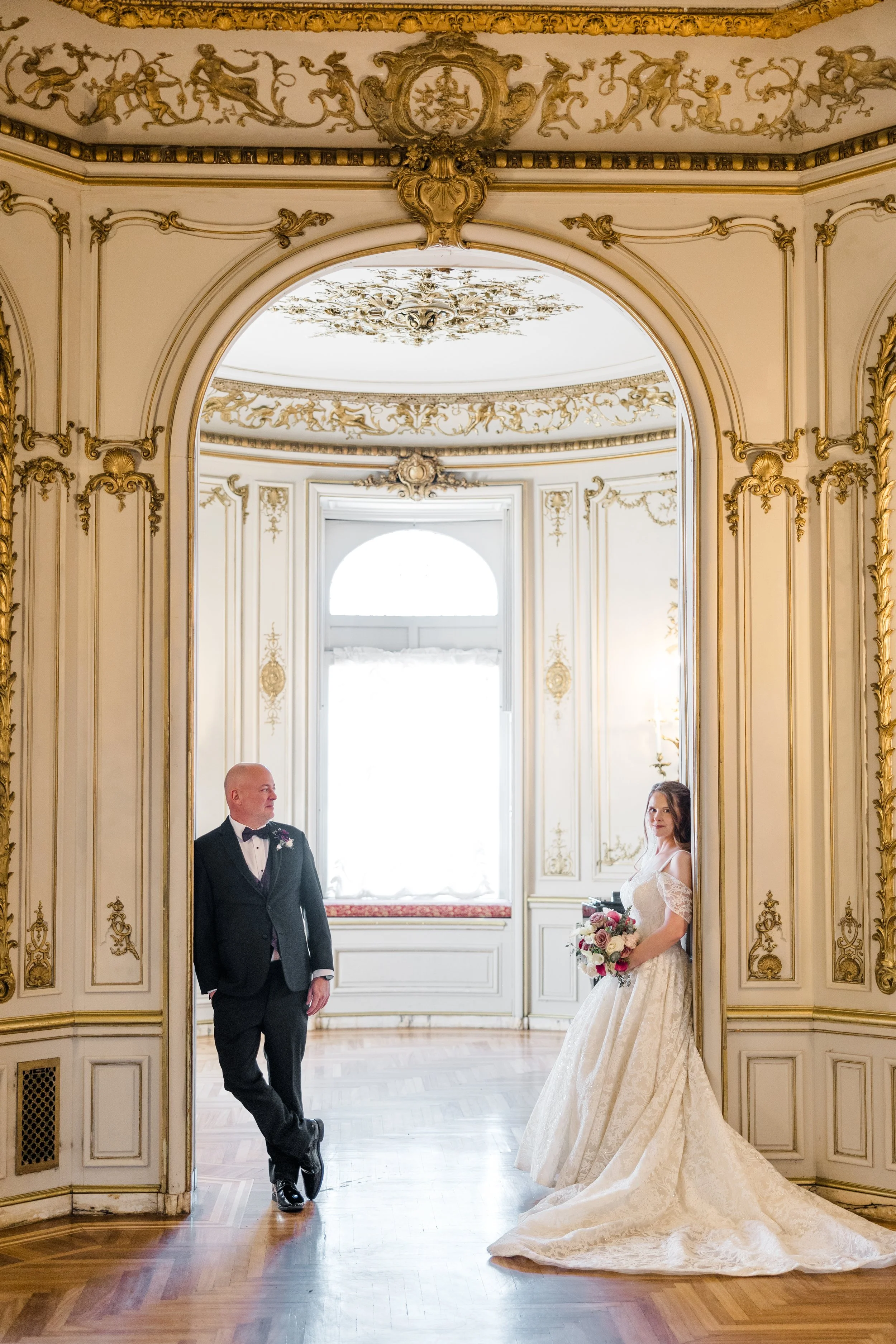 A bride in a white wedding dress holding a bouquet of roses stands near a mirror, while a groom in a black tuxedo leans against the mirror frame inside an ornate, gilded room with elaborate gold and white decoration.