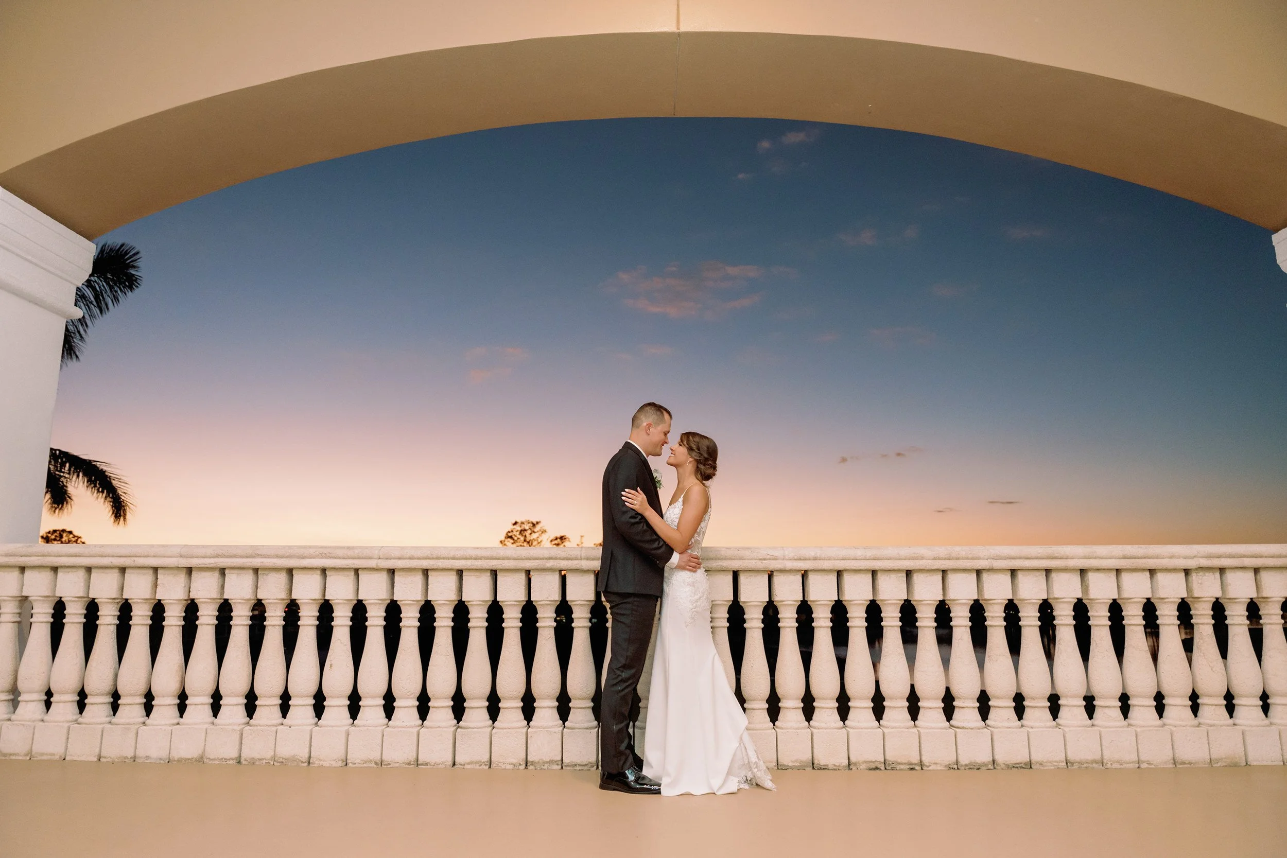 A bride and groom standing close together on a balcony at sunset, gazing at each other, with a sky showing shades of blue and pink and palm trees in the background.