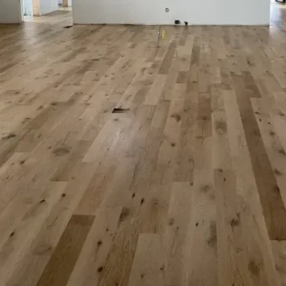 Interior of a house under construction with unfinished walls, recessed lighting, and a newly installed hardwood floor.