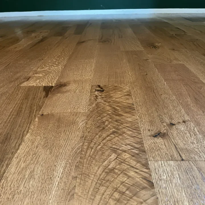 Close-up of a wooden hardwood floor with natural grain and a smooth finish, showing the wall baseboard and green wall in the background.