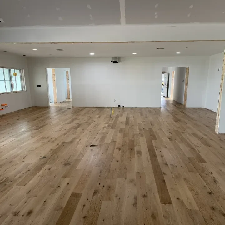 Interior of a house under construction with unfinished walls, recessed lighting, and a newly installed hardwood floor.