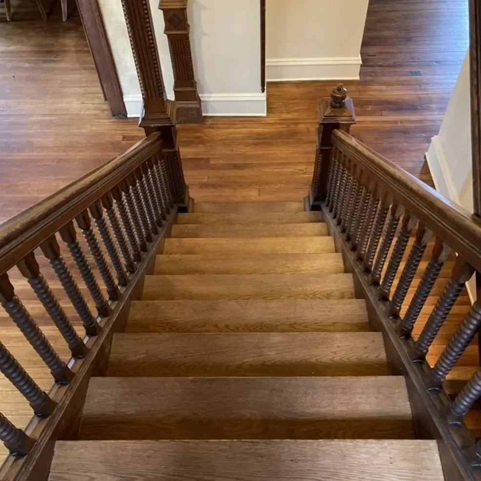 View of a wooden staircase from the top, showing the handrails and spindles, leading down to a landing with hardwood flooring.
