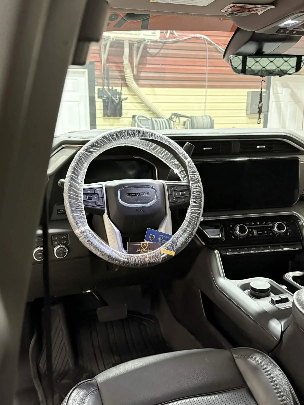 Interior of a vehicle showing a black dashboard, steering wheel wrapped in plastic, digital display, climate controls, and center console, with a view of the garage outside.