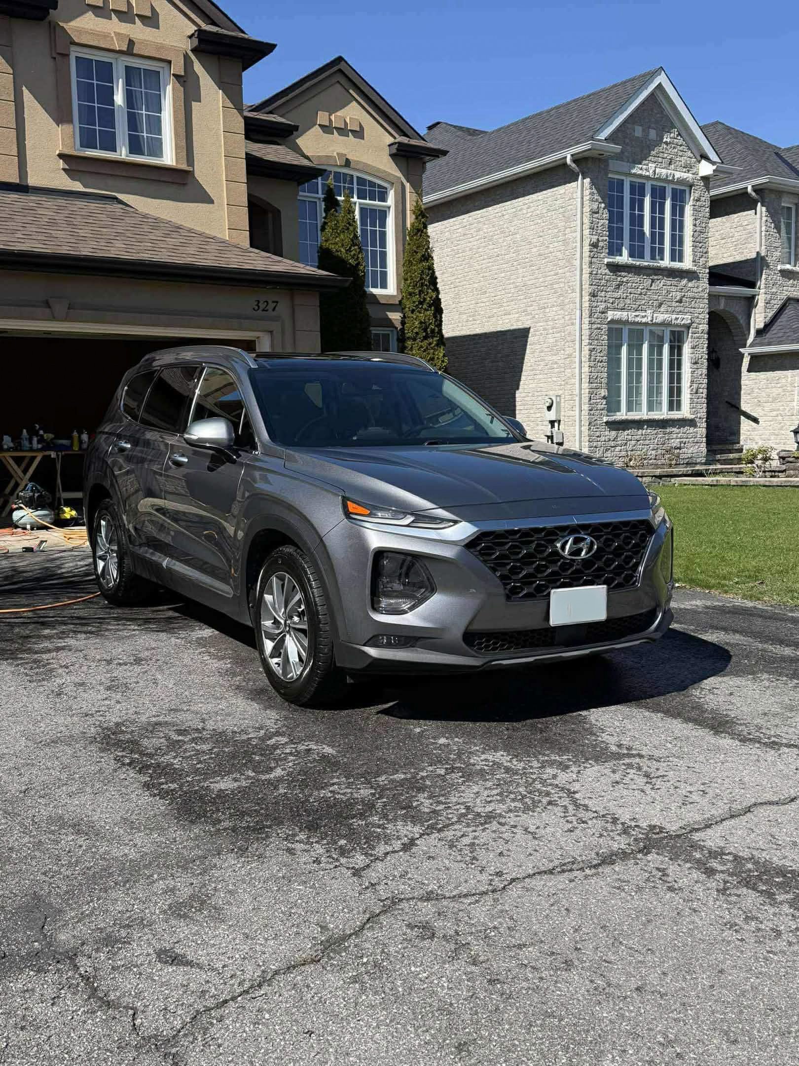 A gray Hyundai SUV parked on a driveway in front of a two-story house with a garage, in a suburban neighborhood under a clear blue sky.