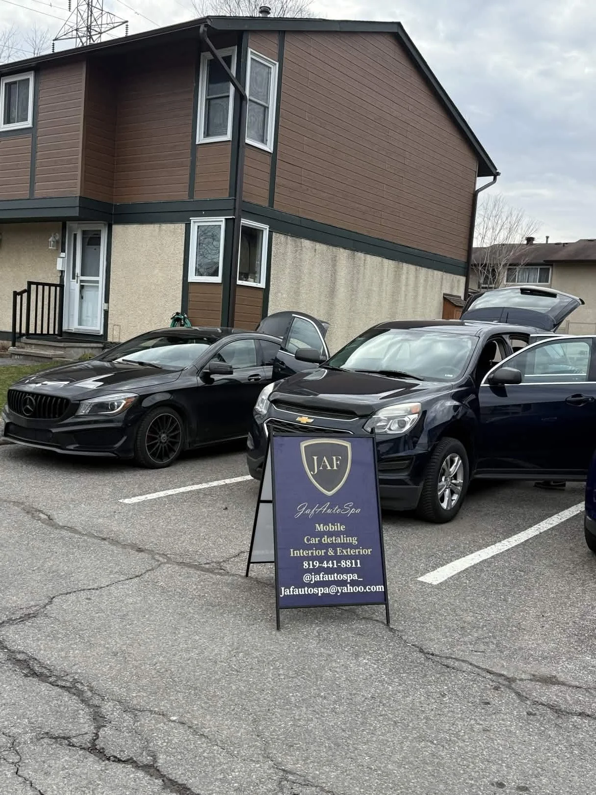 Parking lot with two parked cars in front of a two-story residential building, and a sign advertising a mobile car detailing service.