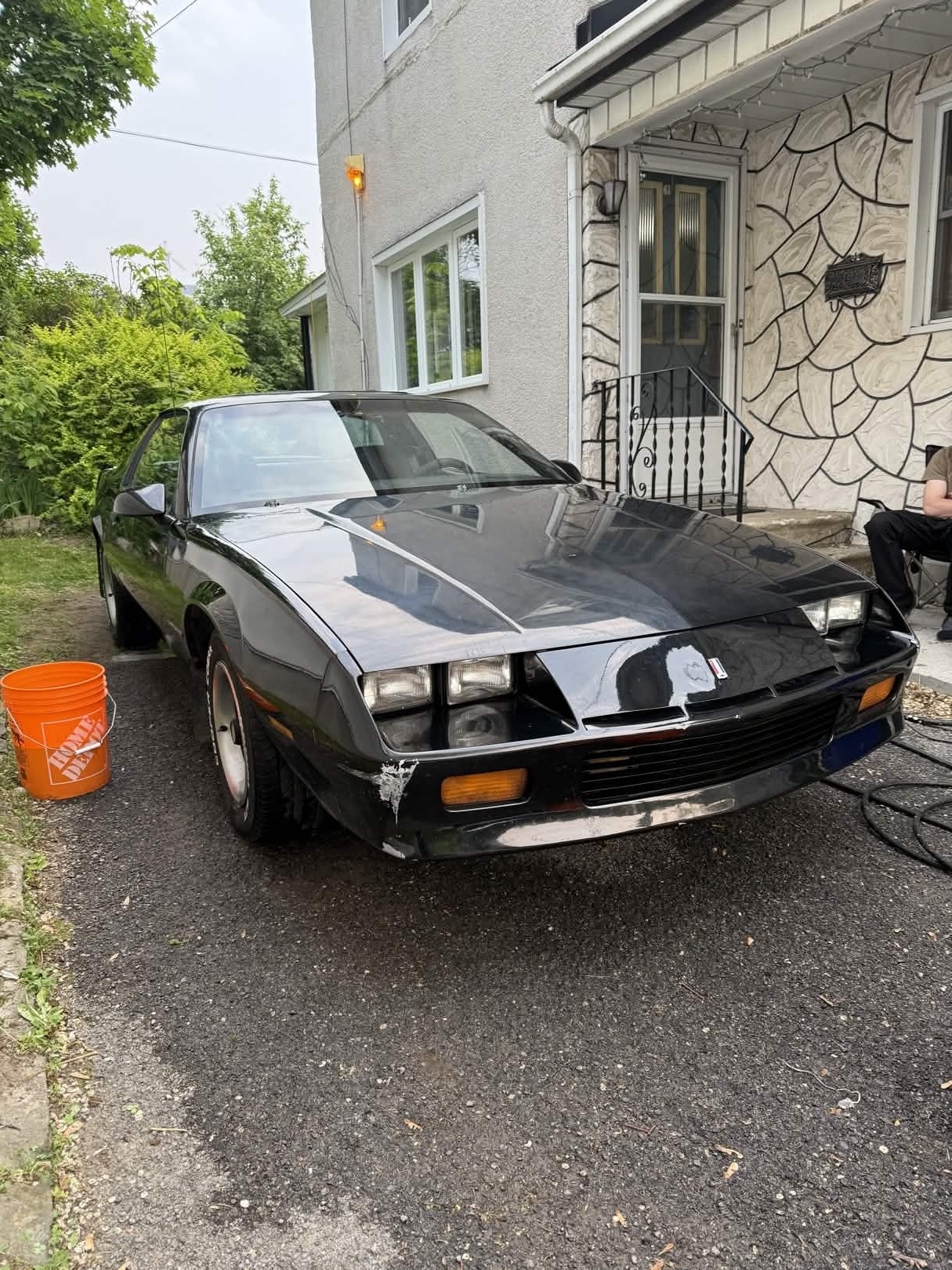 Black classic sports car parked in driveway next to a house with a textured stone and stucco exterior, and a man sitting on the porch.