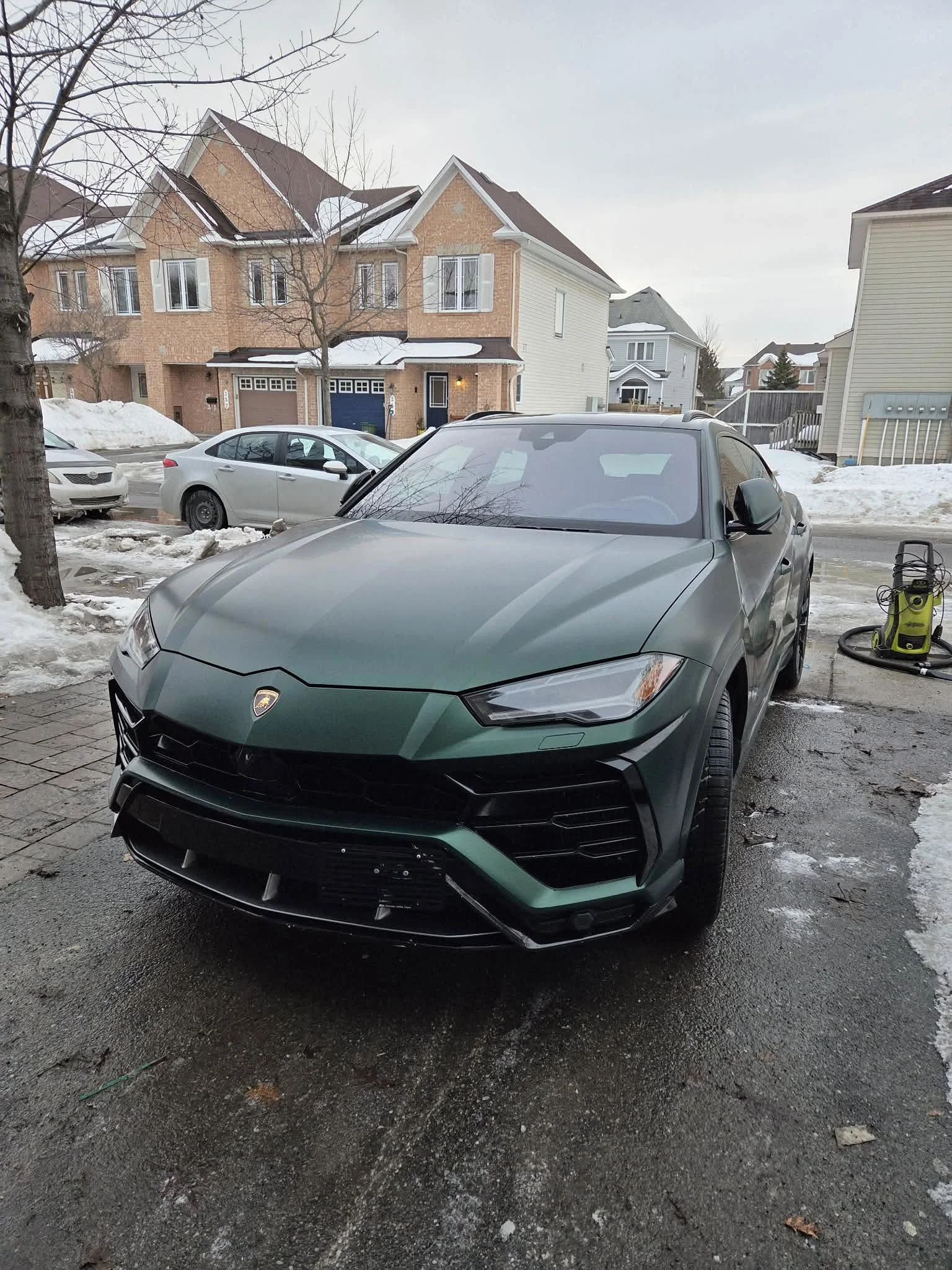 Green Lamborghini Urus parked on a driveway with snow and ice, residential houses in the background, and a pressure washer on the side.