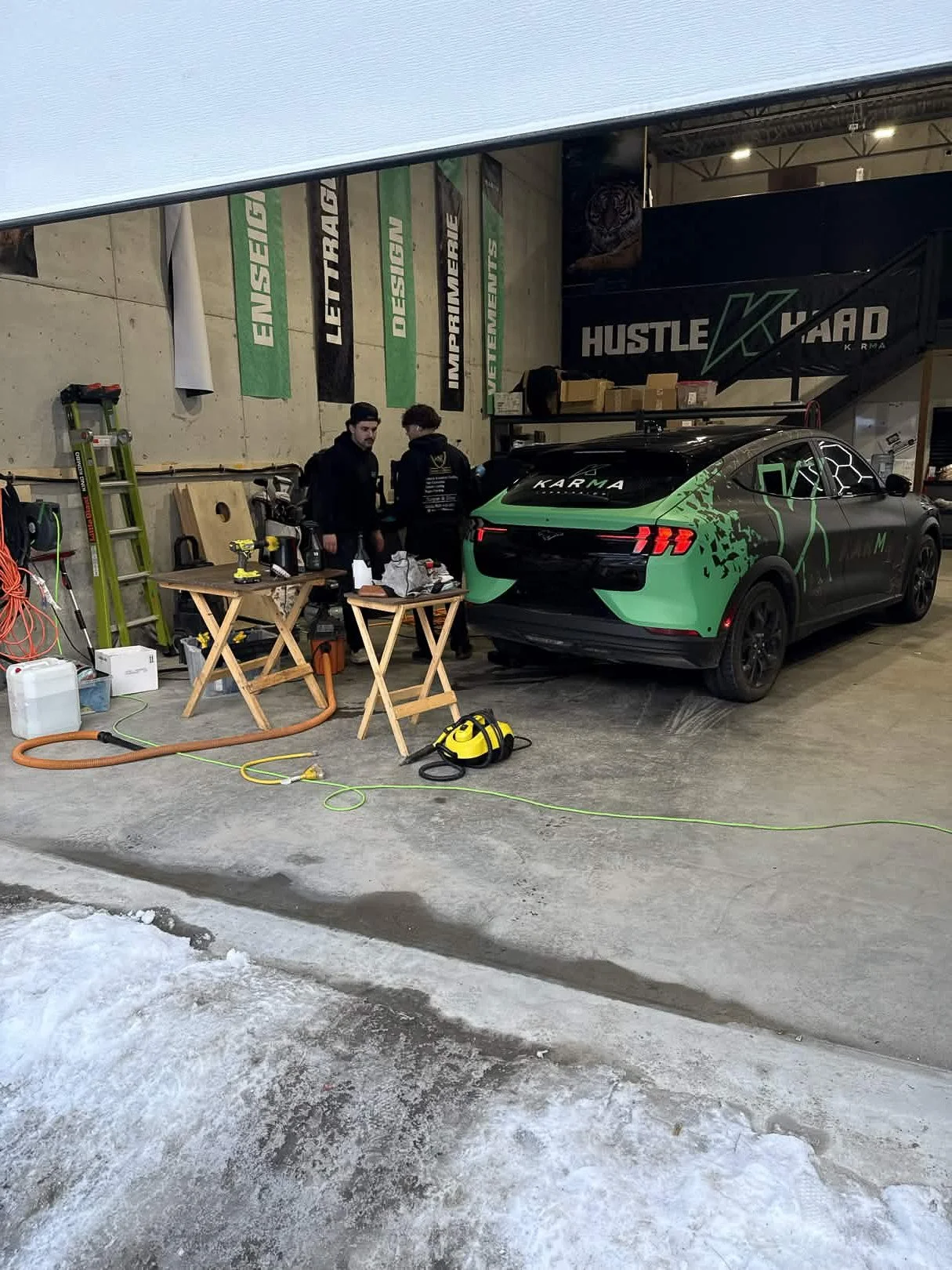 Inside a garage or workshop with a black and green Karma car, two men working on it, tools and equipment on tables, a yellow vacuum, a ladder, and hanging banners with words like 'ENSLIGHT,' 'LET TRAC,' 'DESIGN,' and 'IMPERAME.'