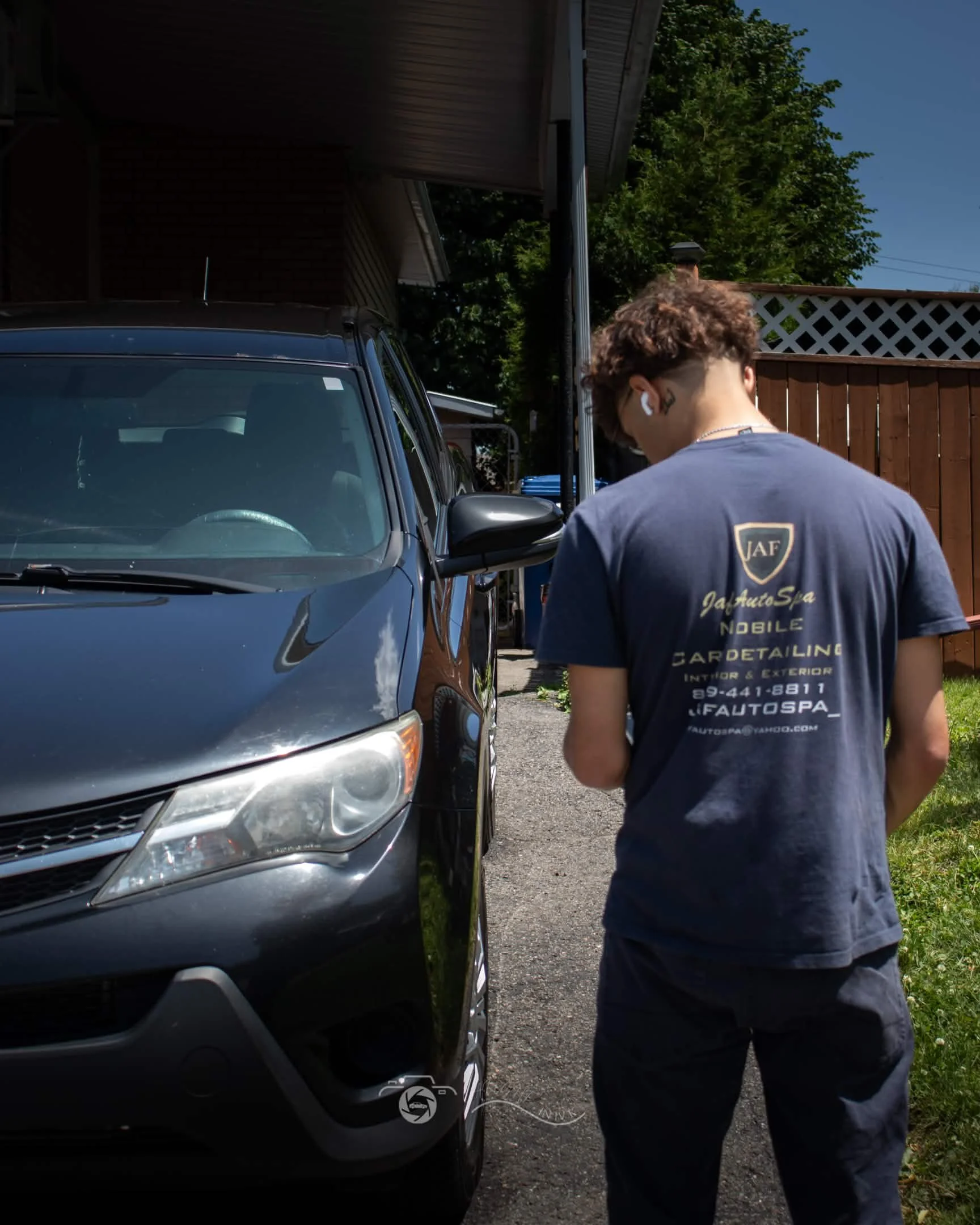A person with curly hair wearing a navy blue T-shirt and black pants stands next to a gray car in a driveway. The person is facing away from the camera, and is wearing wireless earbuds. The yard has green grass and a wooden fence, with a house partia