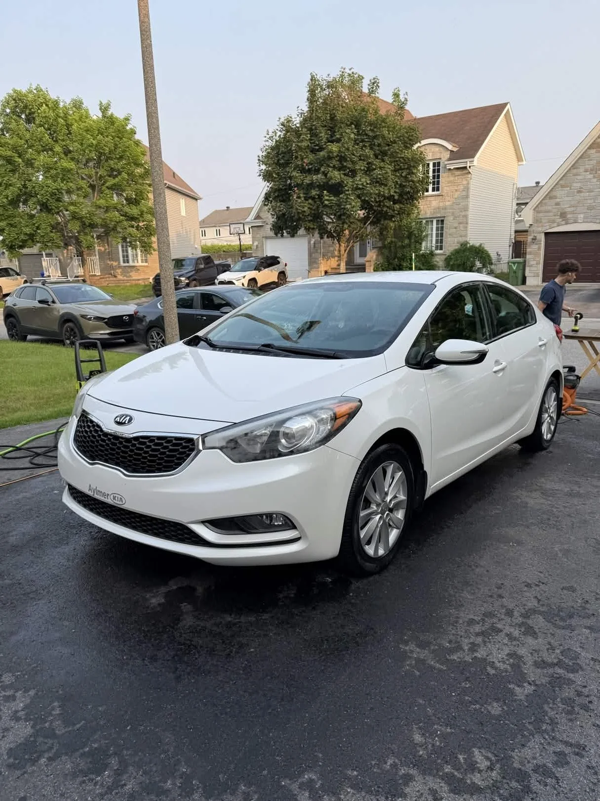 A white Kia sedan parked on a driveway with a house and trees in the background. A person is working at a table nearby.