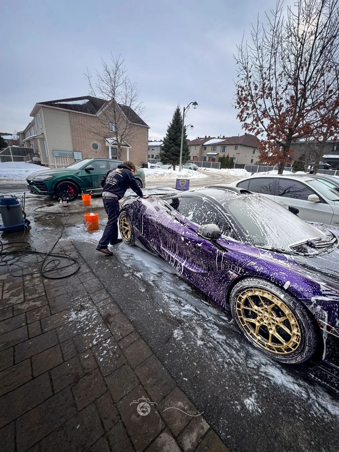 A person washing a purple sports car with a foam brush at an outdoor car wash station in winter, with snow on the ground and other parked cars in the background.