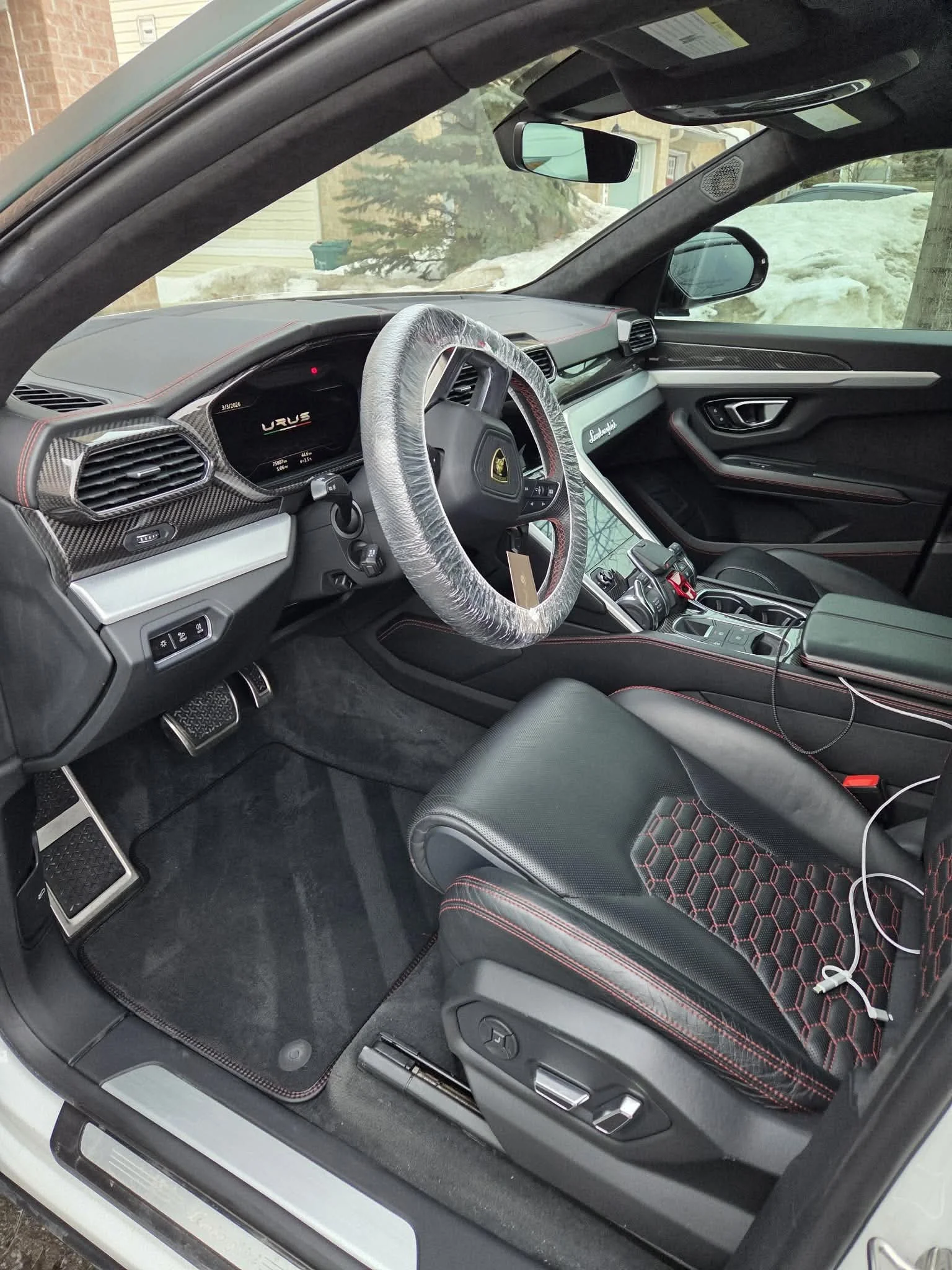 Interior of a luxury sports car with leather seats, digital dashboard, carbon fiber accents, and a steering wheel wrapped in protective plastic.