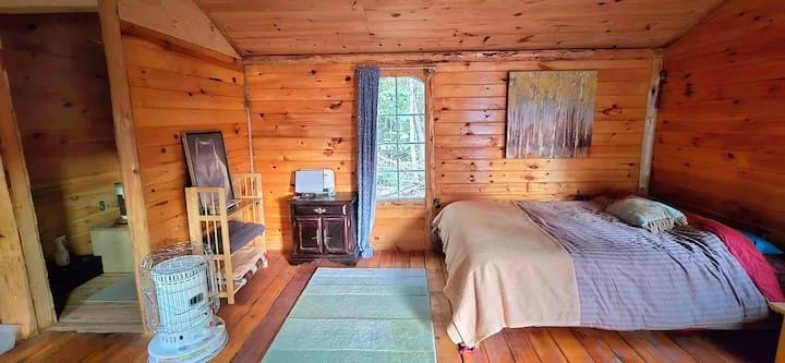 A cozy wooden bedroom with a bed, a window with blue curtains, a small dresser with a mirror, a chair, a painting on the wall, a colorful rug, and a small heater.