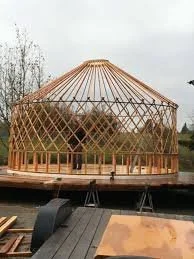 A partially constructed wooden geodesic dome frame outdoors with a cloudy sky in the background. Yurt frame being build and preparing for the yurt raising