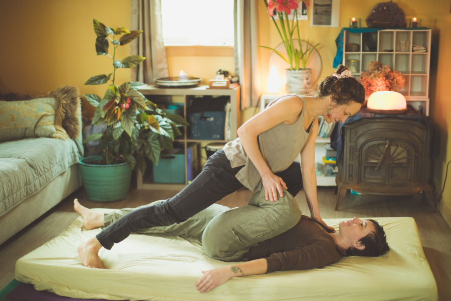 Two women practicing yoga or acro yoga indoors on a bed, with one woman lying on her back and the other woman balancing on her, holding her hips for support.