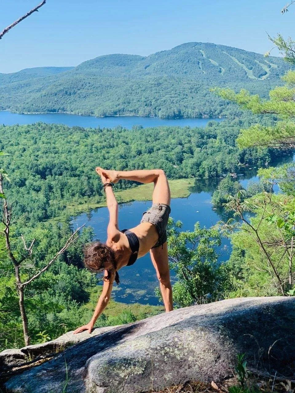 Woman leading and practicing yoga while on a beautiful scenic hike in midcoast maine. A mountain and lake in the background, with a stunning summer scenery.