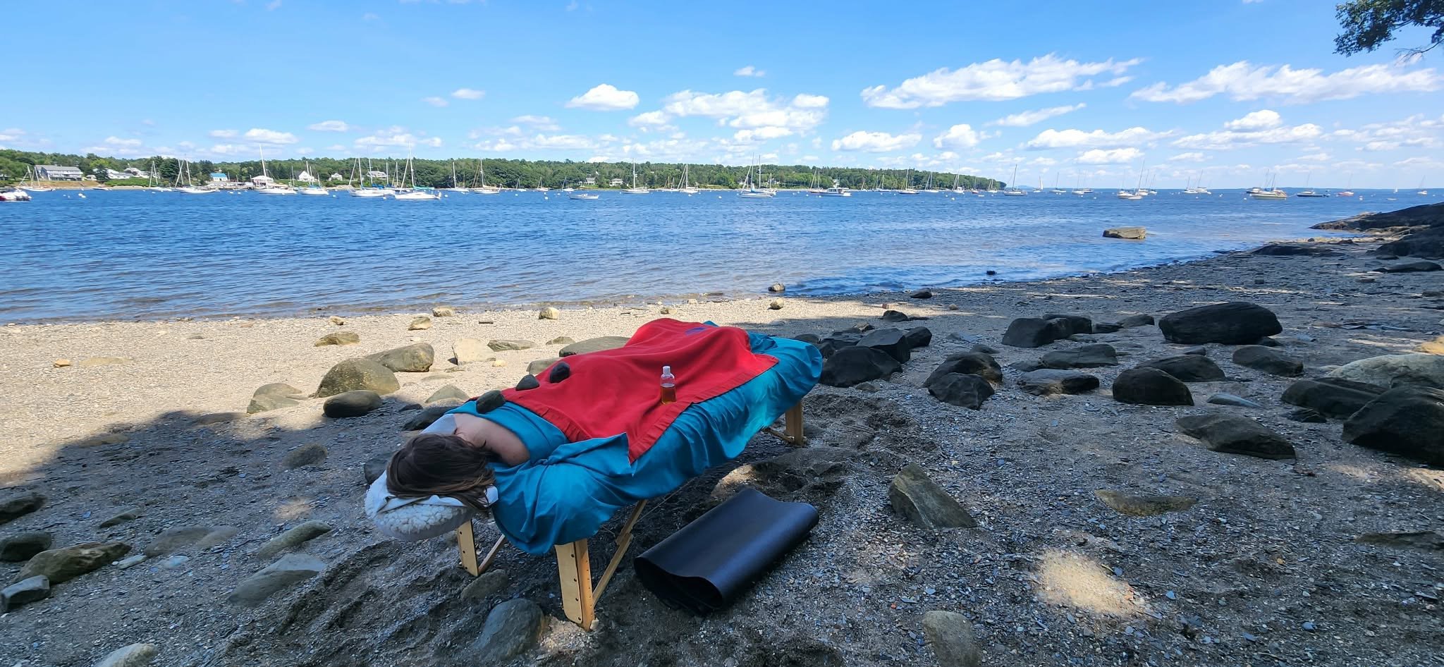 A person receiving a massage, on a beautiful rocky beach in Mid coast Maine. A wonderful light sea breeze with the warmth of summer. Soaking up the ocean view with sailboats passing by in the distance.
