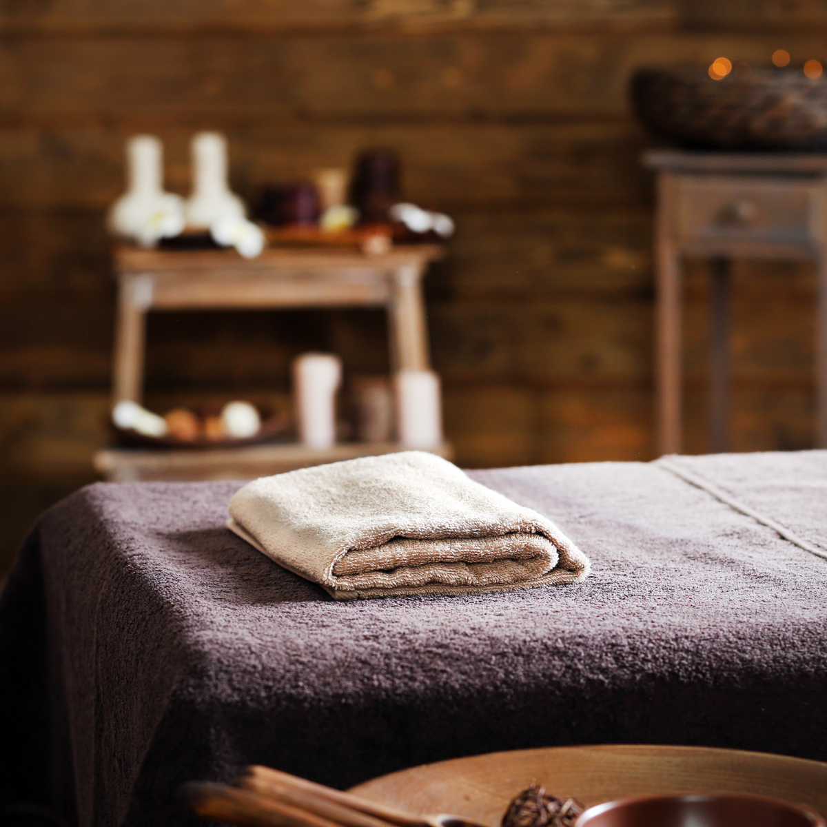 A folded beige towel on a massage table covered with a dark brown blanket, in a rustic wooden spa room.