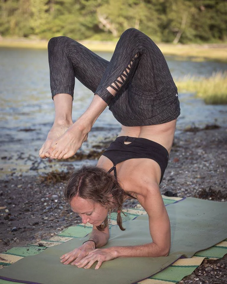 Woman practicing yoga outdoors on a mat by a body of water, balancing on her forearms with legs bent over her back.