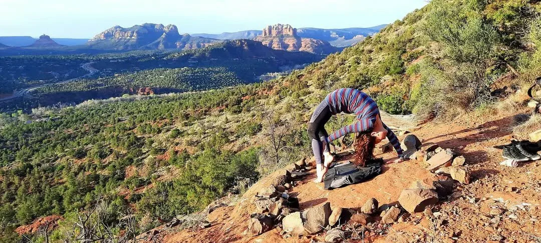 A woman doing a backbend yoga pose on a dirt trail with a scenic view of forested hills and mesas in the background.