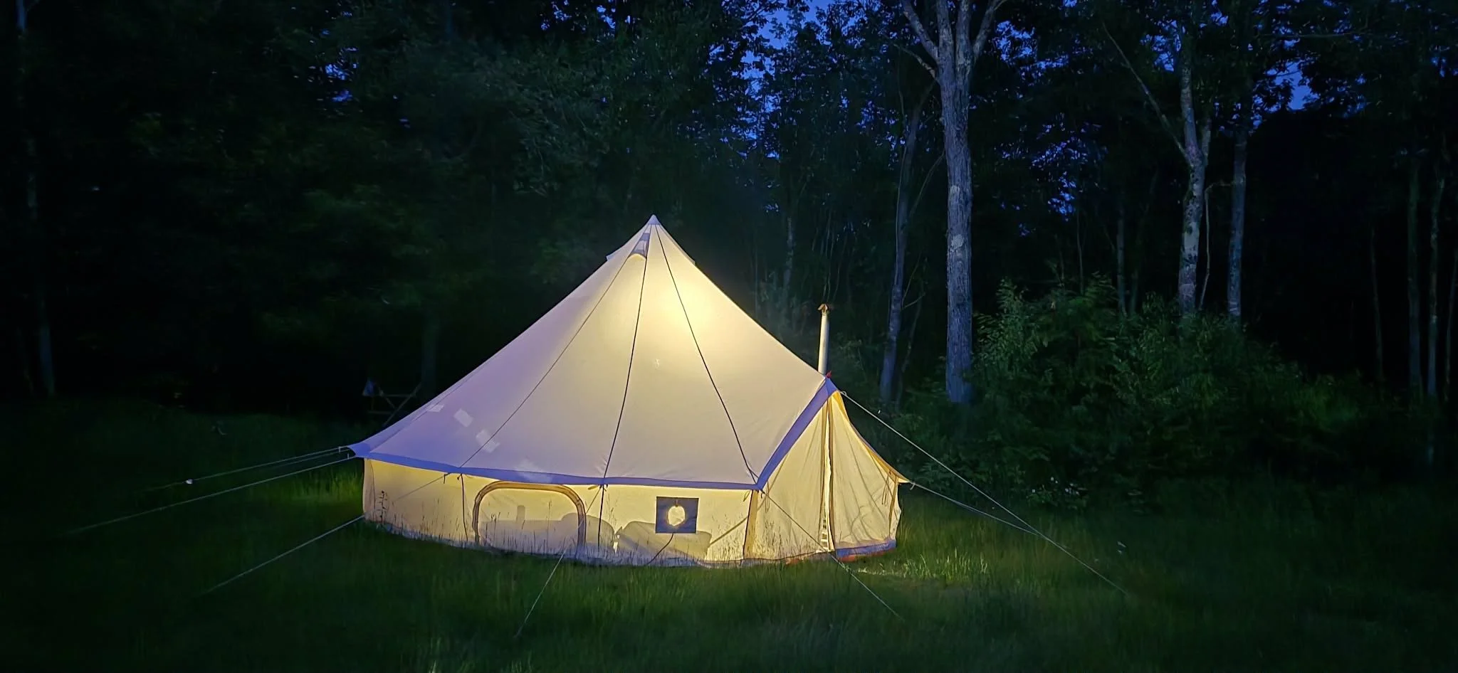 A large illuminated tent pitched on a grassy clearing in a forest at dusk or night.