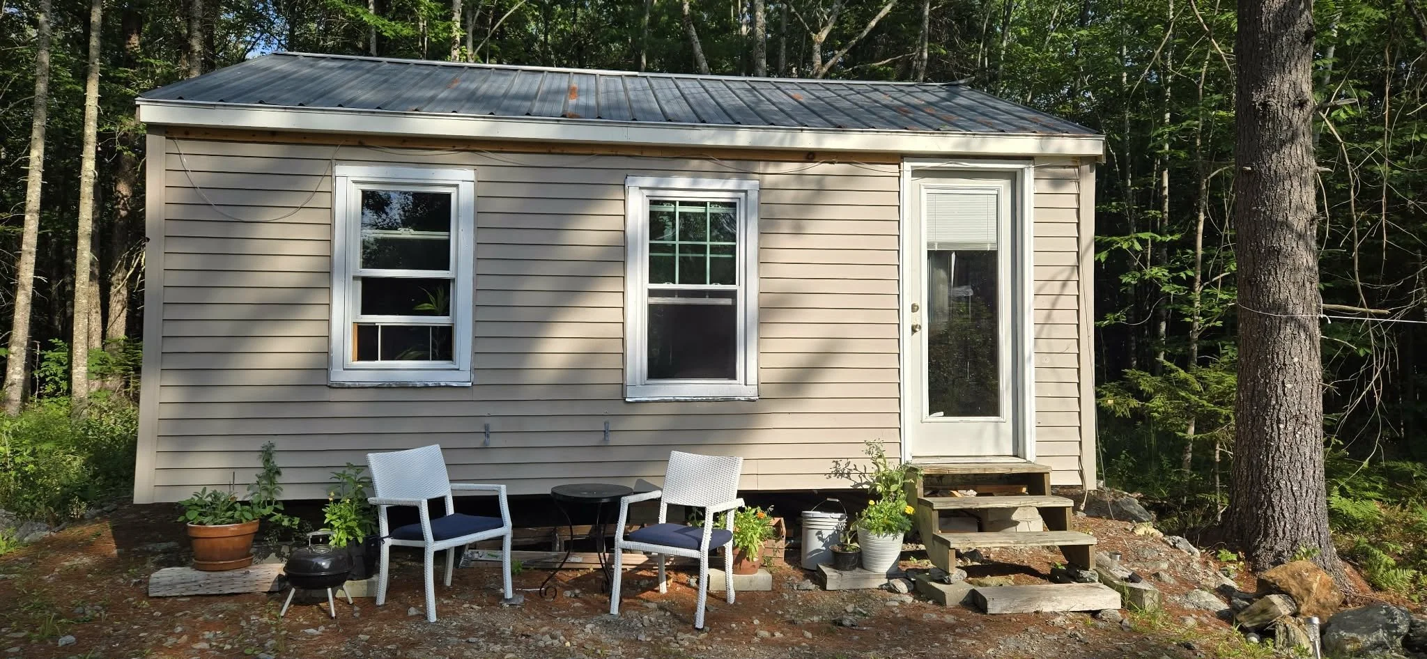 Small beige house with white trim, two windows, and a glass door, set in a wooded area. In front, there is outdoor furniture including two chairs, a small table, and potted plants.