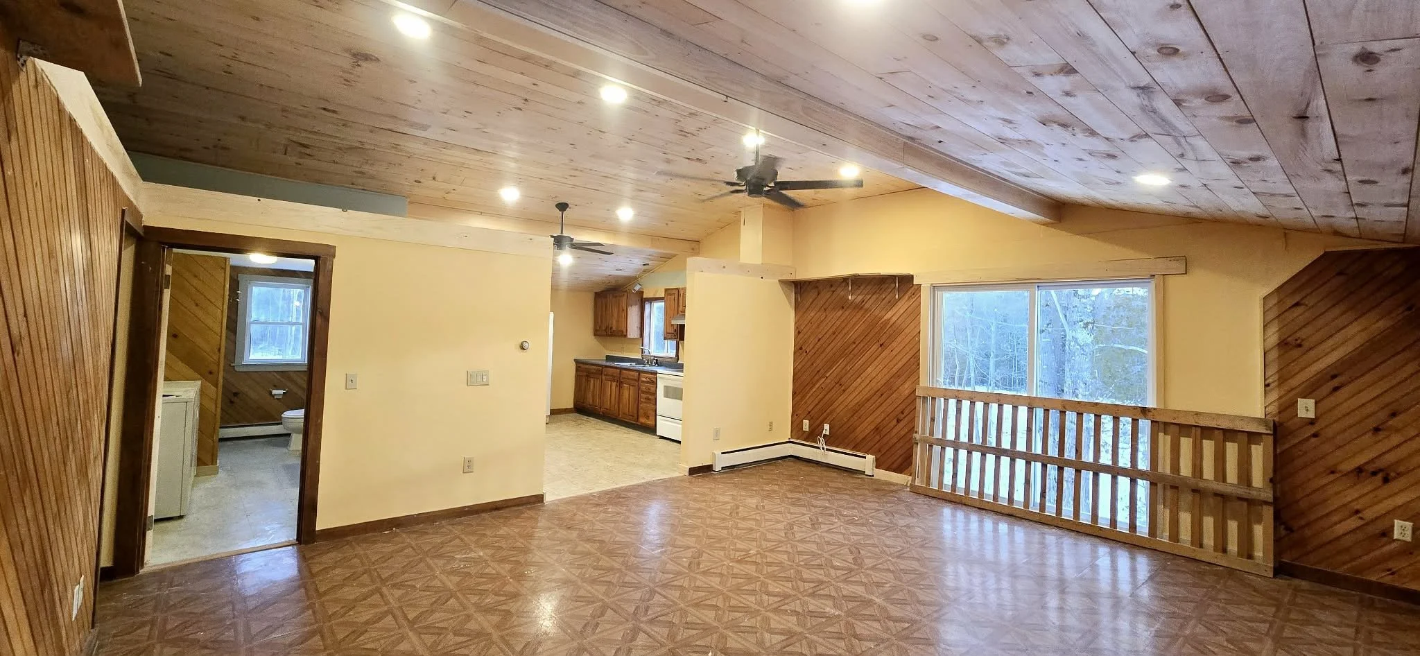 Empty living room with wooden floors, wood-paneled walls, and ceiling, a large window with a view of trees, ceiling fans, and a small railing near the window. Adjacent kitchen area with wooden cabinets and white appliances, and a bathroom visible through a doorway.