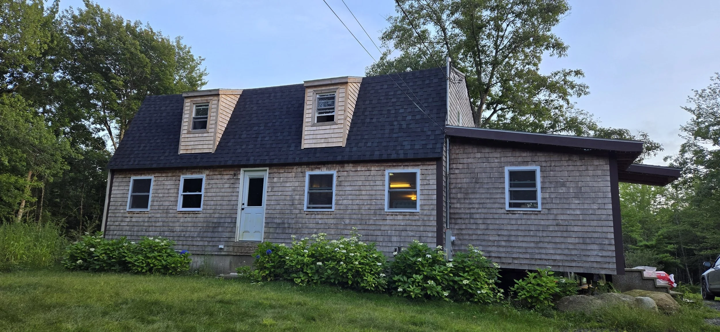 A two-story house with wooden shingles, multiple windows, a door, and dormer windows on the roof, surrounded by greenery and trees.