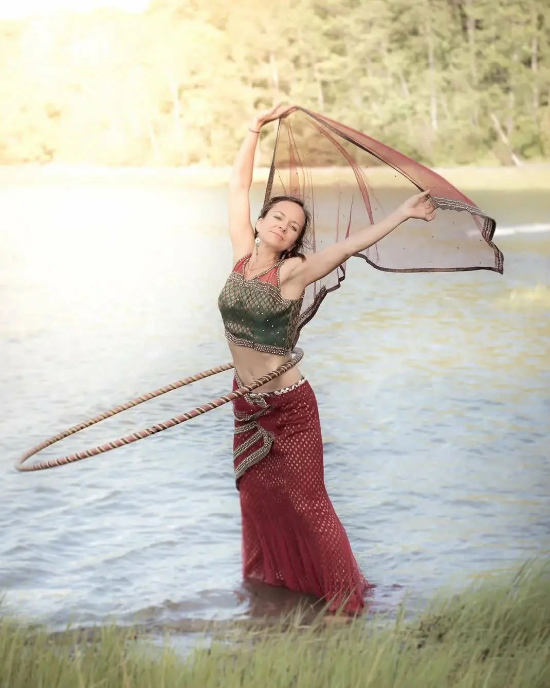 Woman in traditional attire standing in water, holding a parasol and a hoop, with trees and a lake in the background.
