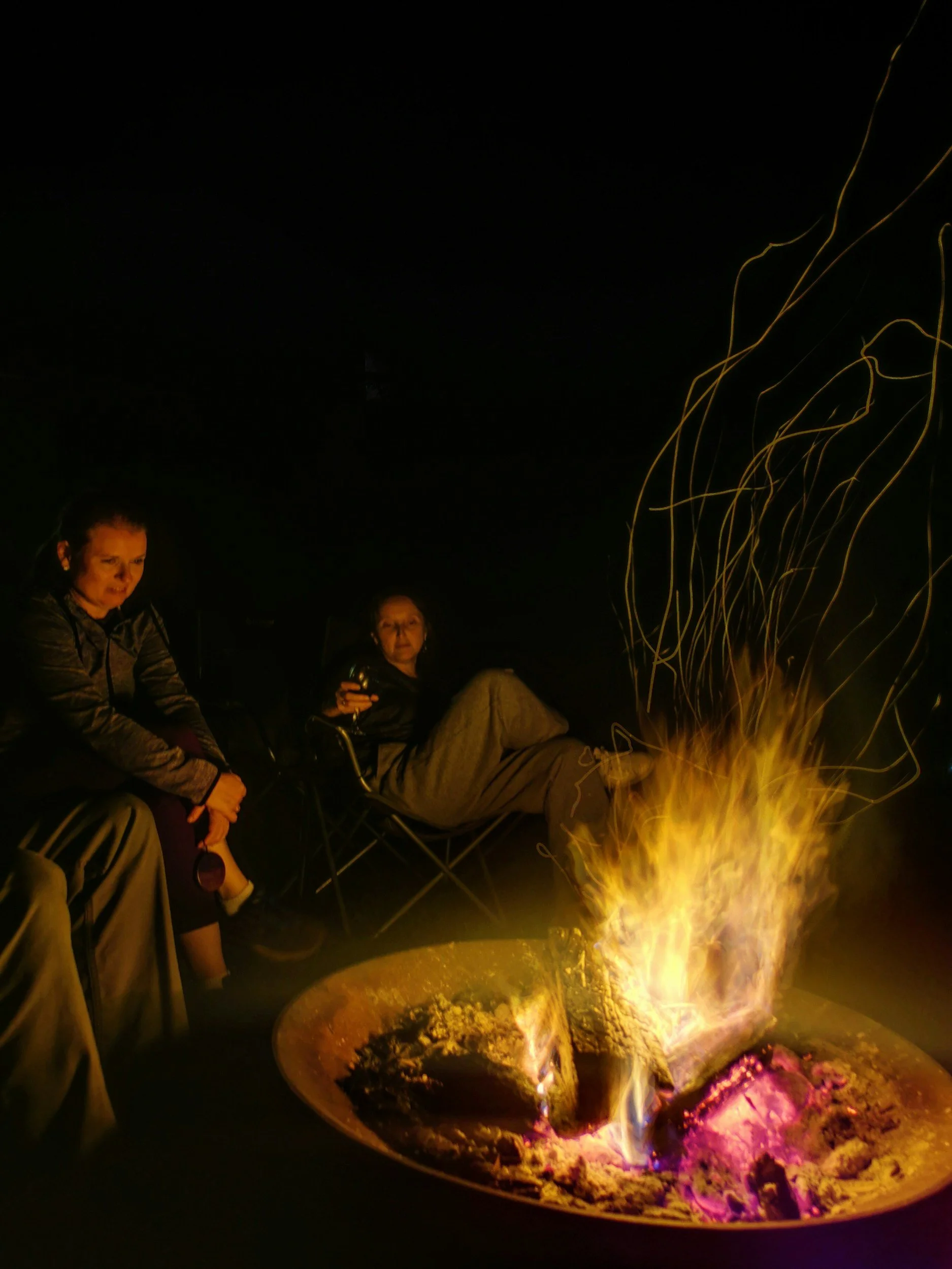 People sitting near a campfire at night, with glowing embers and sparks flying.