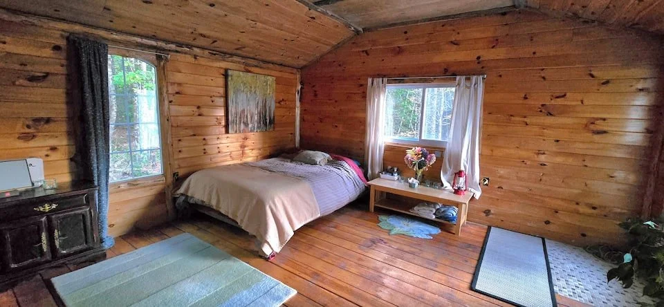 A cozy bedroom with wooden walls and floors, featuring a bed with beige and pink bedding, a small wooden table with flowers and decorative items, a window with white curtains, and a dresser. There is a blue and gray striped rug near the bed and a small plant in the corner.