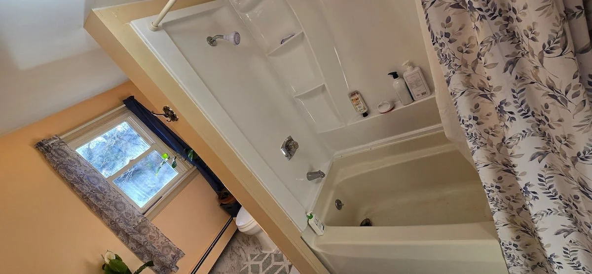 A bathroom with a bathtub and shower, a window with lace and dark curtains, and floral-patterned bedspread on a nearby bed.