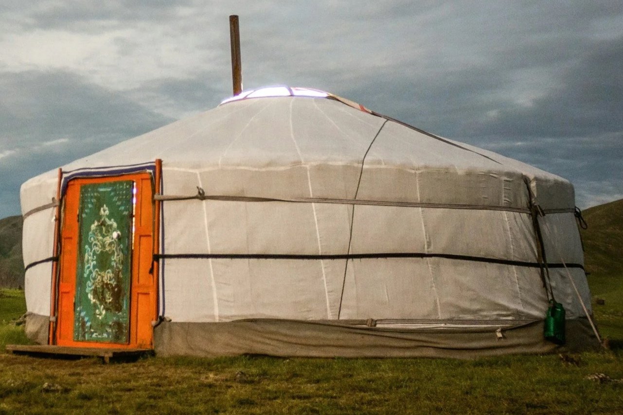 A round, white yurt with a colorful ornate door set on green grass under a cloudy sky.