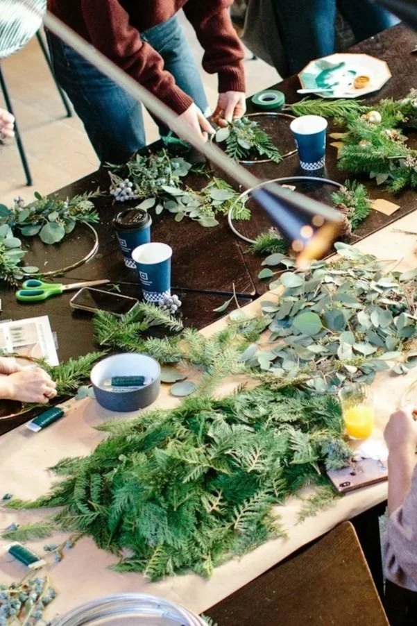 People making Christmas wreaths with evergreen branches and foliage on a table, surrounded by scissors, cups, and other craft supplies.