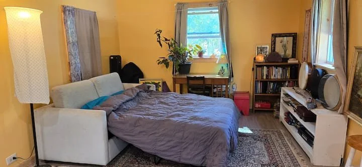 A bedroom with a bed, a window, a bookshelf, and various decor items, illuminated by natural and artificial light.