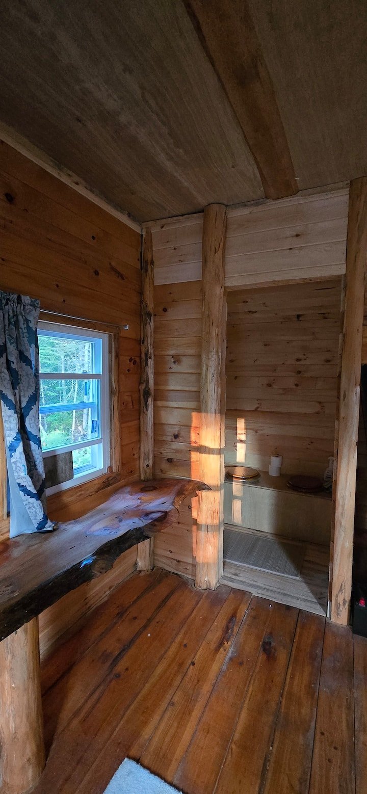 Interior of a rustic wooden cabin room with a window, wooden walls, a small wooden bench, and a countertop with a toilet paper roll.