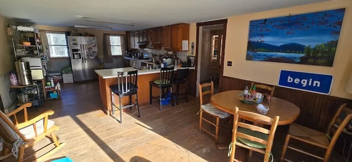 Open kitchen and dining area with wooden furniture, a round dining table with chairs, a kitchen island, and kitchen appliances. There is a large landscape painting on the wall and a blue sign that says 'begin'.