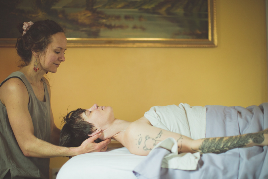 Woman providing a massage to a young woman lying on a massage table in a room with yellow walls and a landscape painting