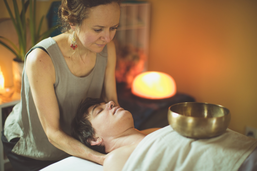 A woman receiving a sound healing or massage therapy in a calming, dimly lit room with warm lighting, a Himalayan salt lamp, and a brass singing bowl on her chest.