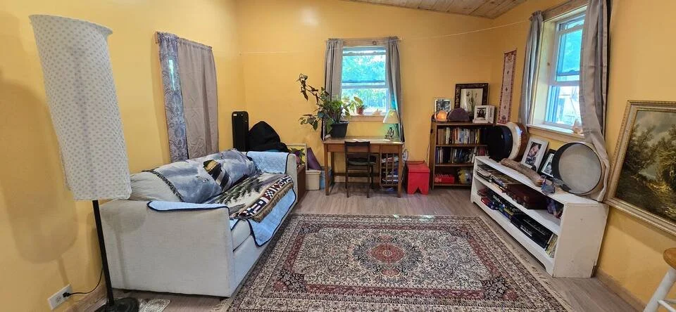A living room with yellow walls, two windows with blue curtains, a white sofa with blankets, a tall white lamp, a coffee table with a plant, a bookshelf with photos and records, a rug with a pattern, and framed pictures on the walls.