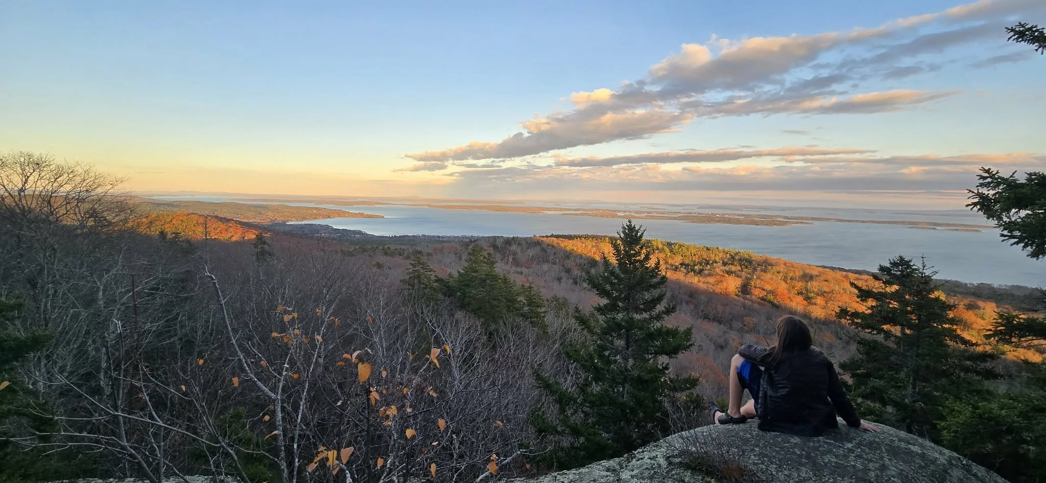 A person sitting on a large rock overlooking a scenic landscape of mid coast, rolling hills covered with trees in autumn colors, under a partly cloudy sky during sunset.