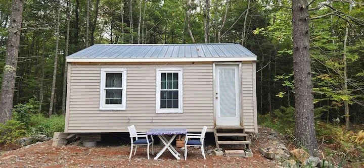 Small beige house with metal roof, three windows, a white door, and a small outdoor table with two chairs, situated in a wooded area.
