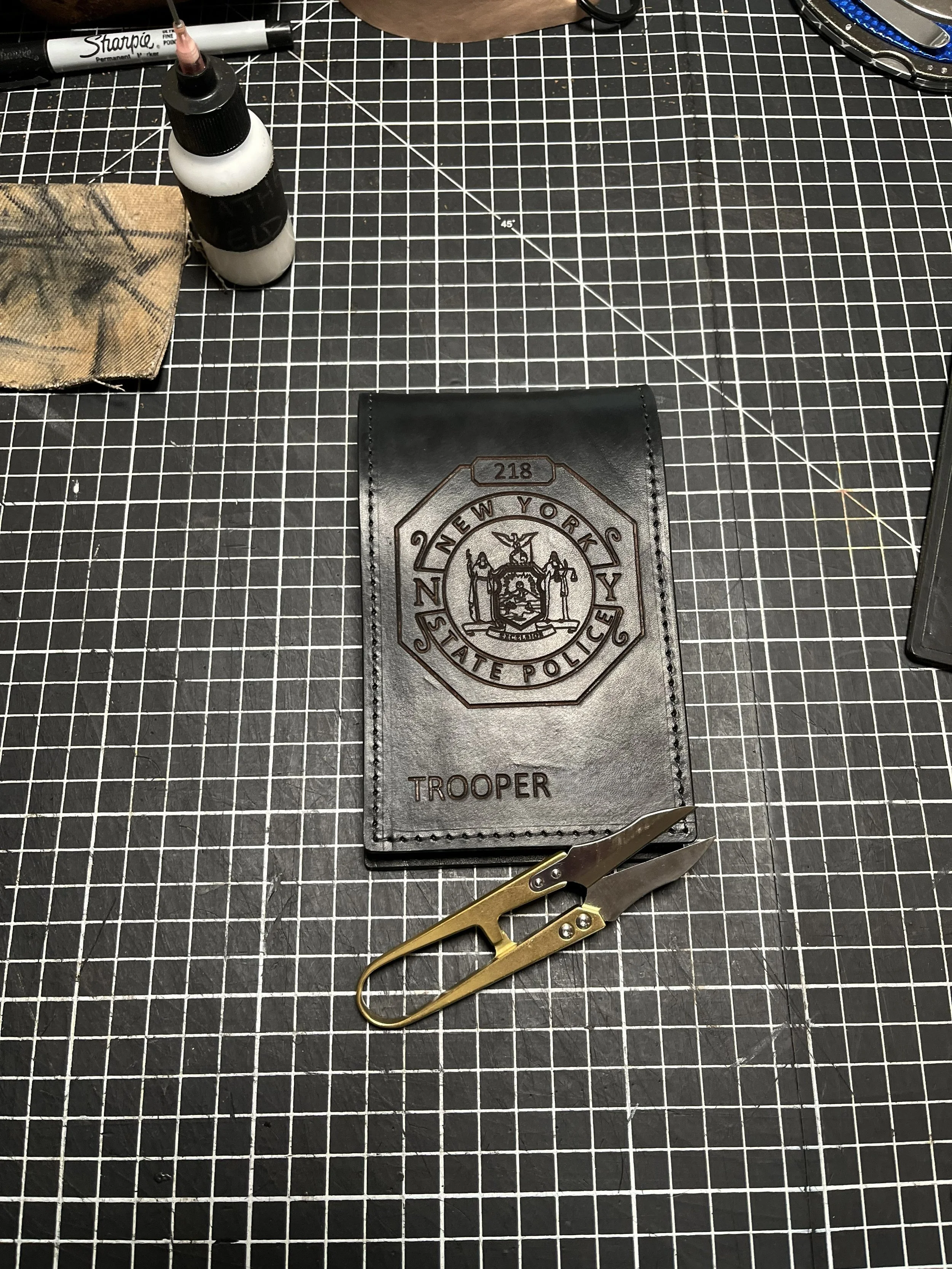 A black work badge with embossed lettering and an emblem on it, labeled 'Trooper' at the bottom, placed on a black cutting mat with measurement grid lines. There is a pair of gold-colored scissors beneath the badge. Additional items on the workspace 