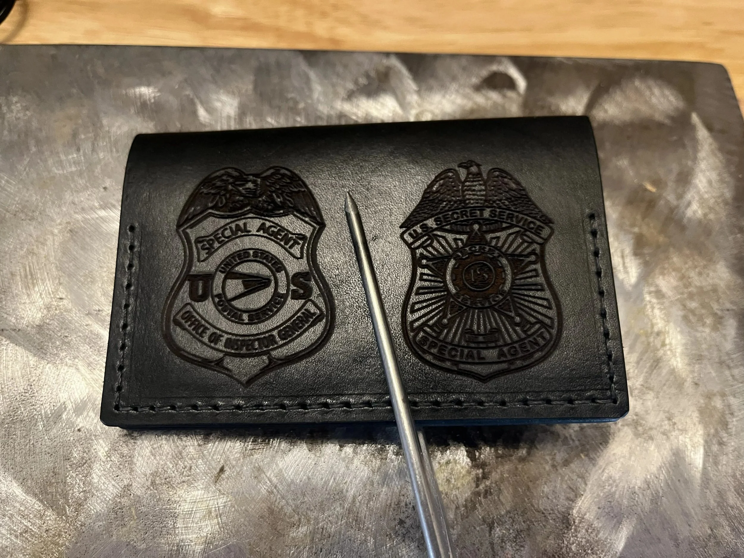 A black leather folder with patches from the United States Secret Service and the Office of Firefighter Crisis, placed on a metallic surface. A tool or pointer is resting on the folder.