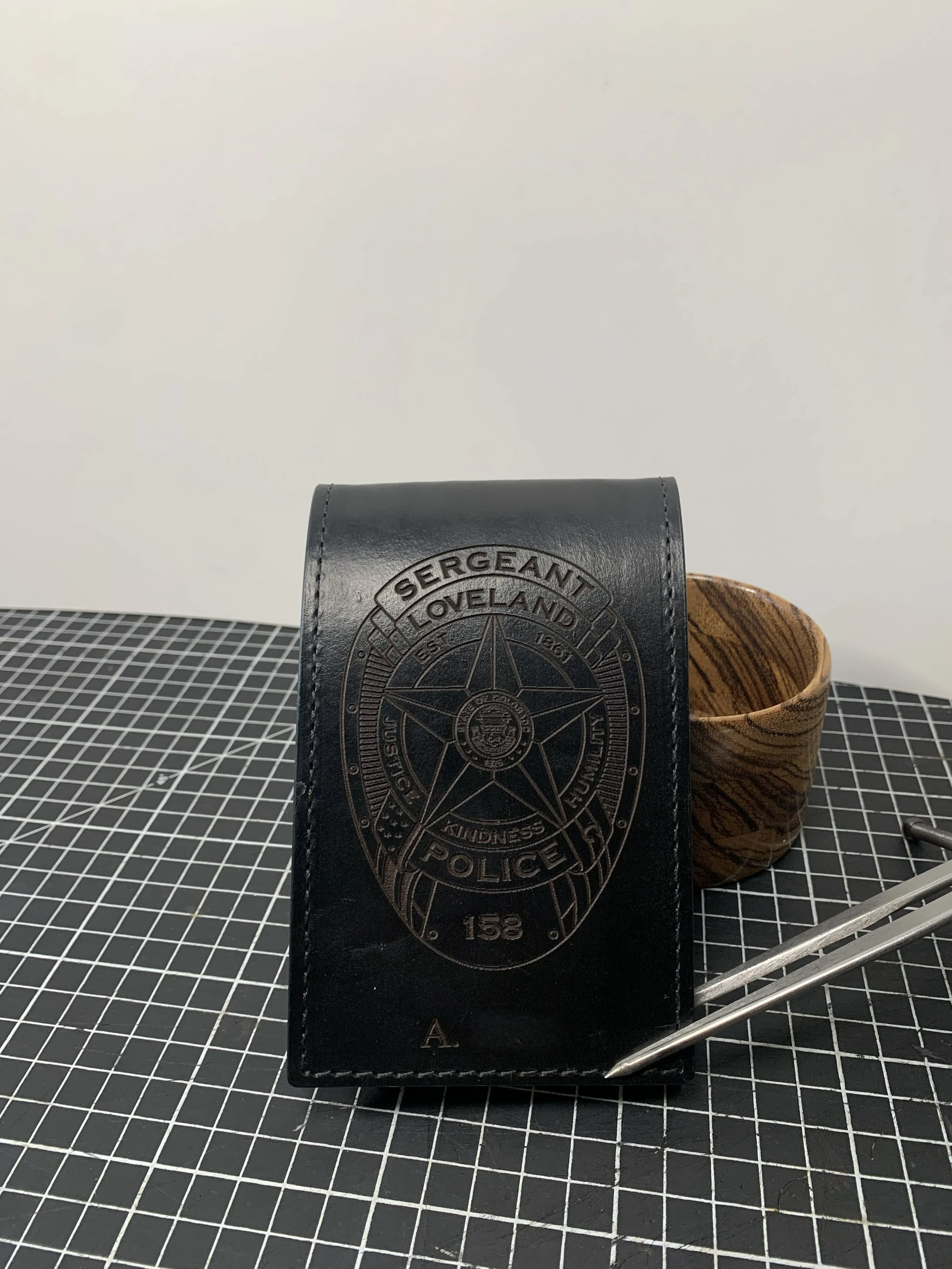 Black police badge wallet with a sheriff's badge emblem, next to a wooden cup and a metal tool on a black and white grid surface with a plain white background.