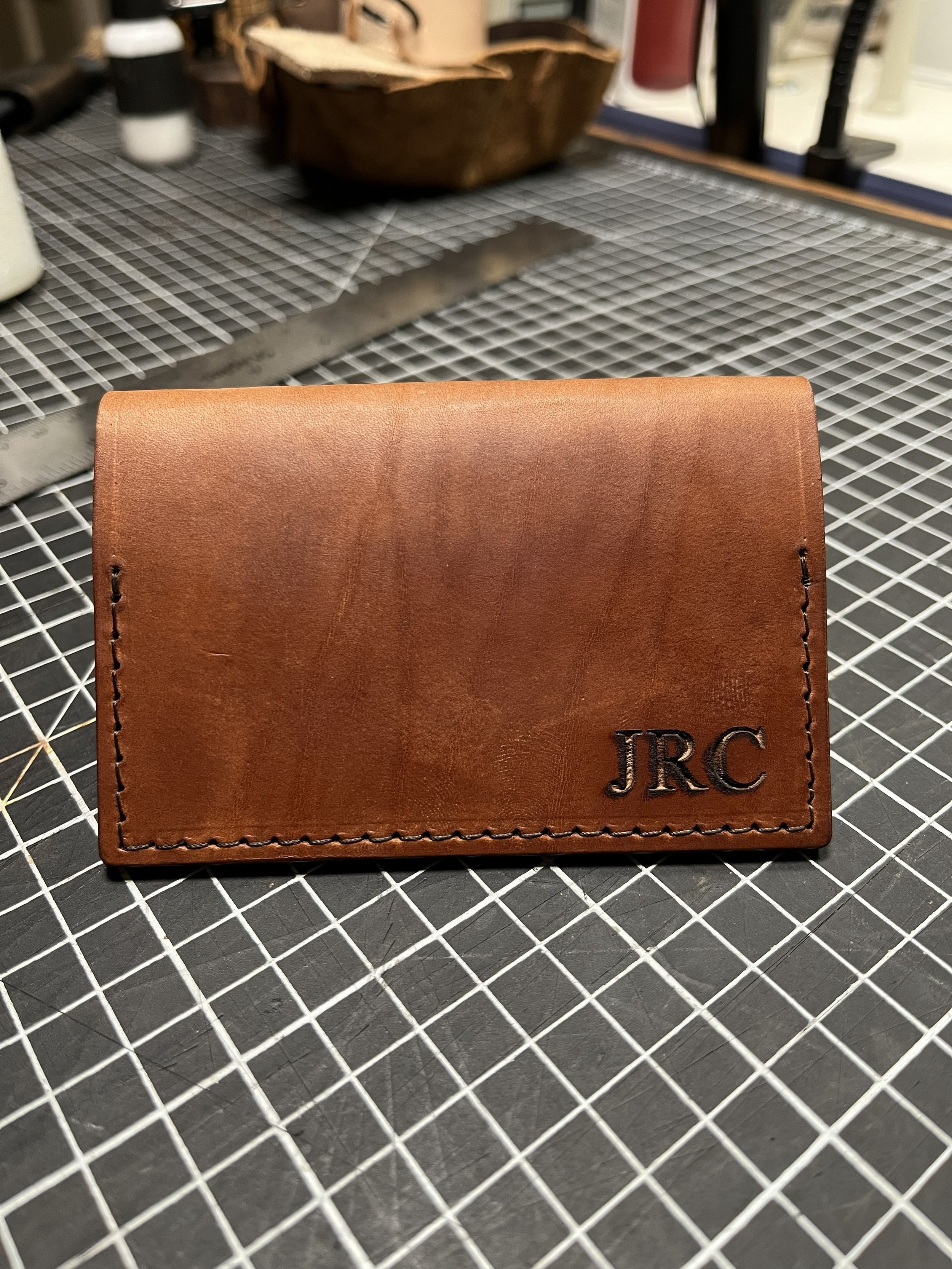 A brown leather wallet with the initials 'JRC' embossed in black on the lower right corner, placed on a black and white grid-patterned surface.