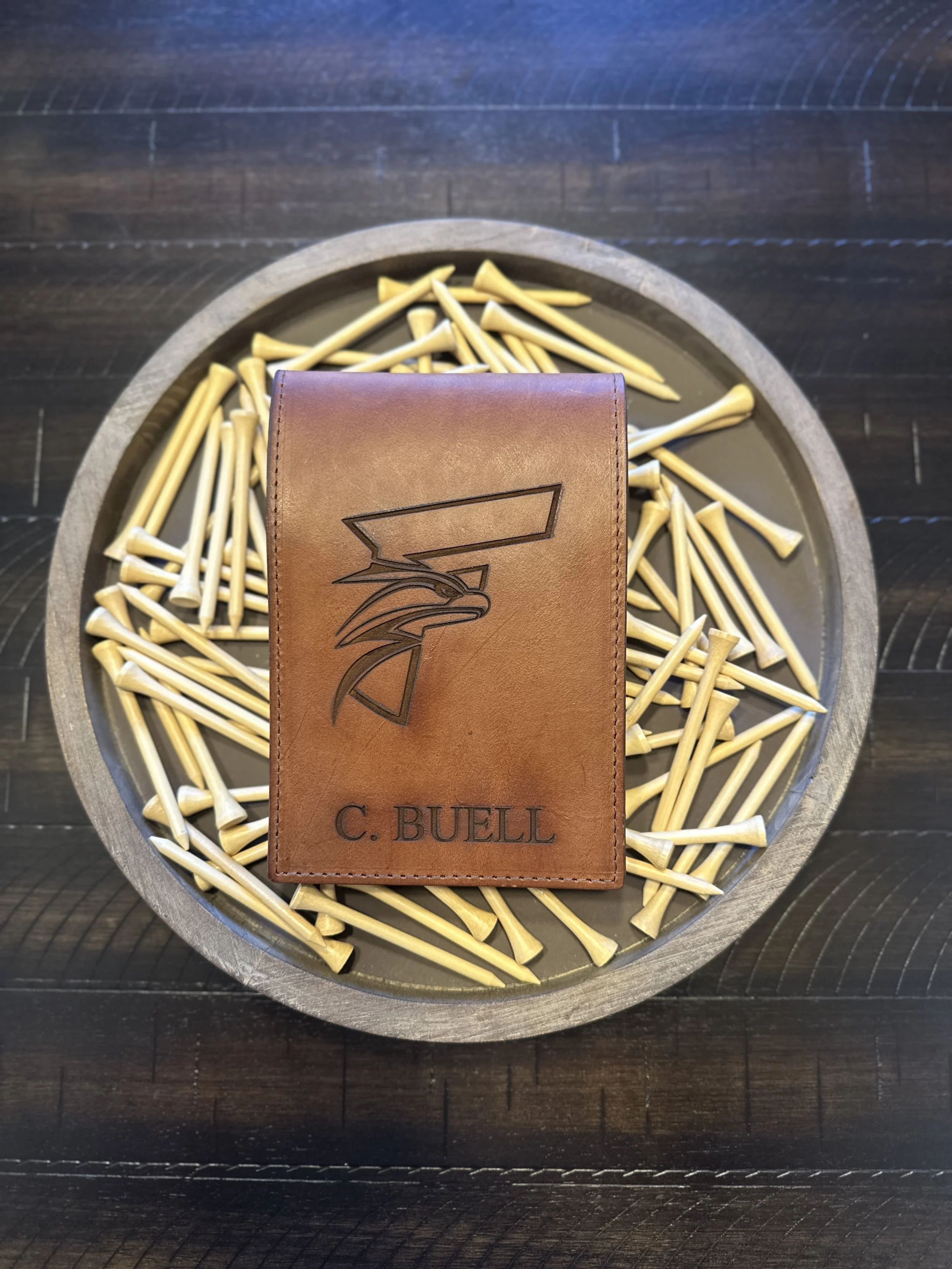 A leather card with the name 'C. BUELL' and a stylized logo, placed on a bed of small yellow bamboo swords inside a circular metal container, on a dark wooden surface.