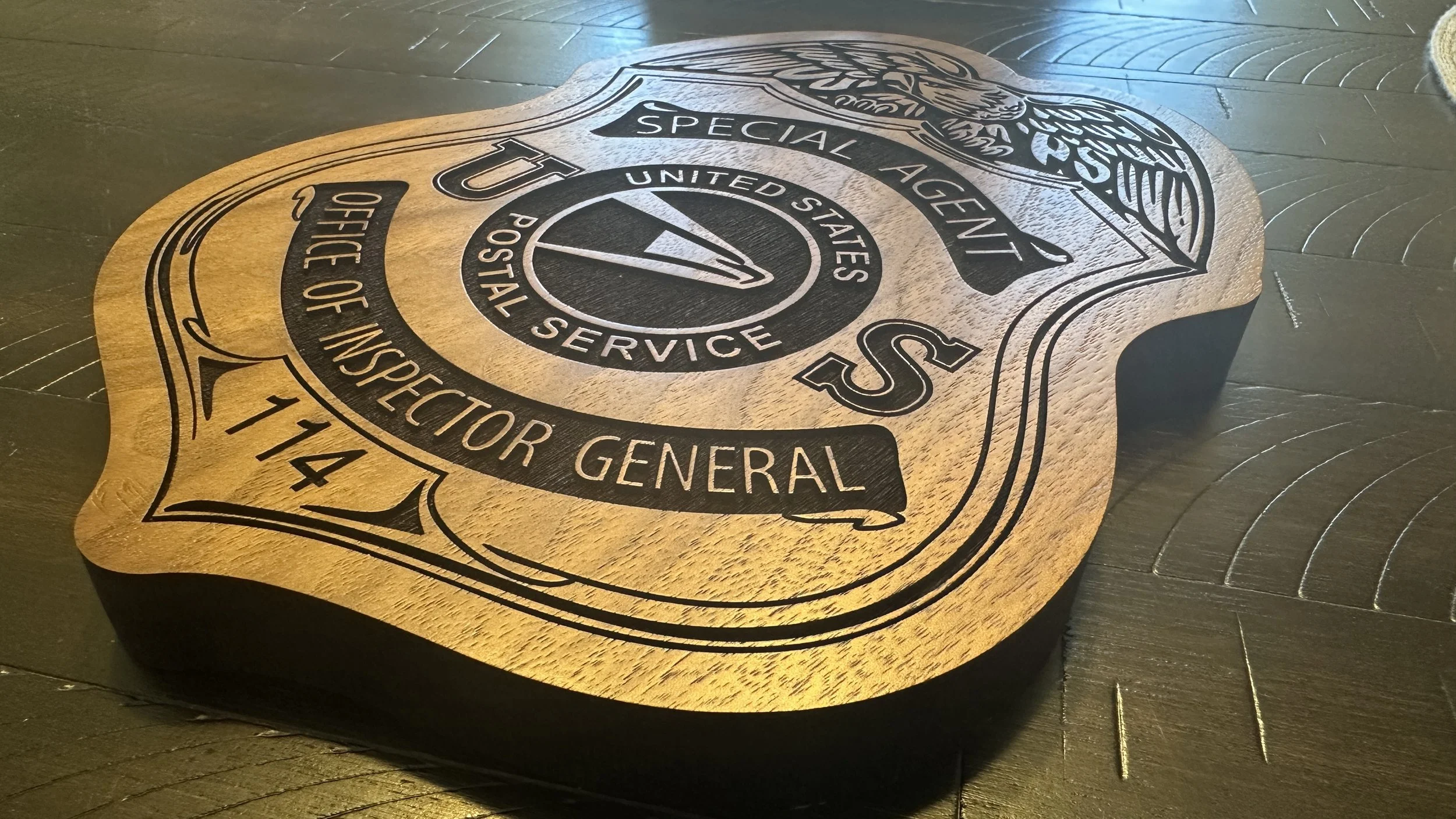 Wooden plaque with a sheriff badge design, labeled 'United States Postal Service, Office of Inspector General, Special Agent, T114' on a dark wooden surface.